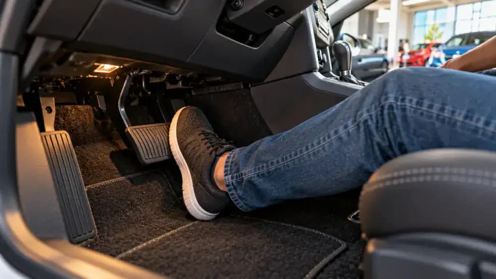 Driver pressing the brake pedal inside a parked American hatchback while the dashboard displays a staged instruction to depress the brake before starting.