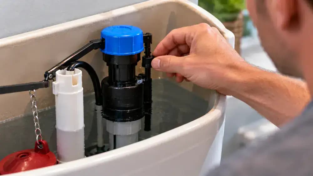 Close-up of a homeowner using a small screwdriver to adjust the float mechanism on a modern toilet fill valve, with the tank water-level mark and vertical overflow tube visible nearby.