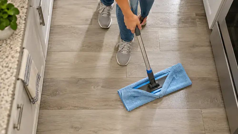 Person drying slightly damp vinyl plank flooring in a residential kitchen with a white microfiber towel attached to a flat mop, carefully removing moisture near plank seams and cabinet edges while a yellow wet-floor warning sign stands at the entrance.