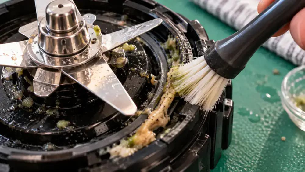 Macro close-up of a narrow soft-bristle brush removing trapped food residue from the circular gasket groove of a detached blender blade housing secured on a dark green silicone mat, with sharp metal blades facing away.
