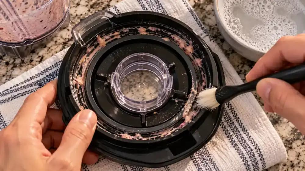 Overhead close-up of an adult cleaning dried strawberry smoothie residue from the narrow grooves of a black blender lid using a soft-bristle brush, with the removable center cap resting separately on a blue towel over a marble countertop.