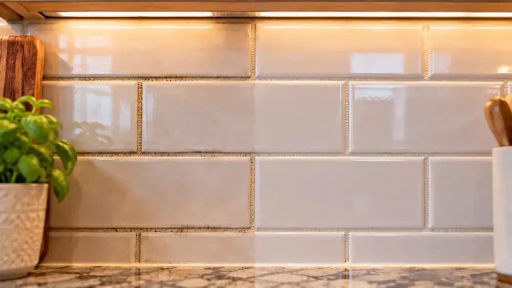 Homeowner wearing blue nitrile gloves inspecting completely dry light gray grout between cream ceramic kitchen backsplash tiles under bright LED lighting, holding a white inspection card beside a darker grout patch for comparison.