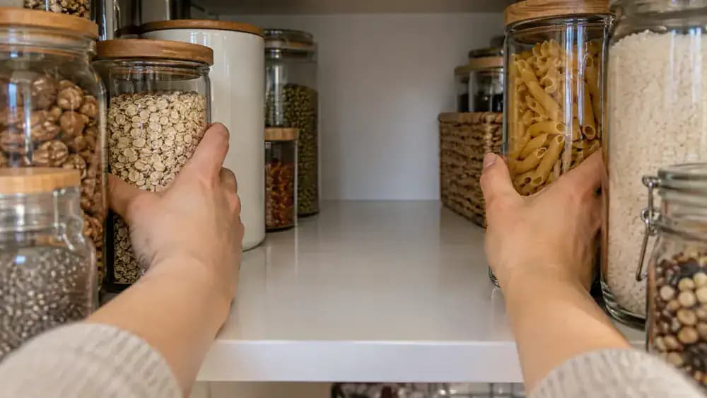Hands adjusting sealed glass jars and pantry containers on a modern kitchen shelf, creating small gaps between jars, cereal boxes, and canisters for better visibility of the dry shelf surface and rear corners.